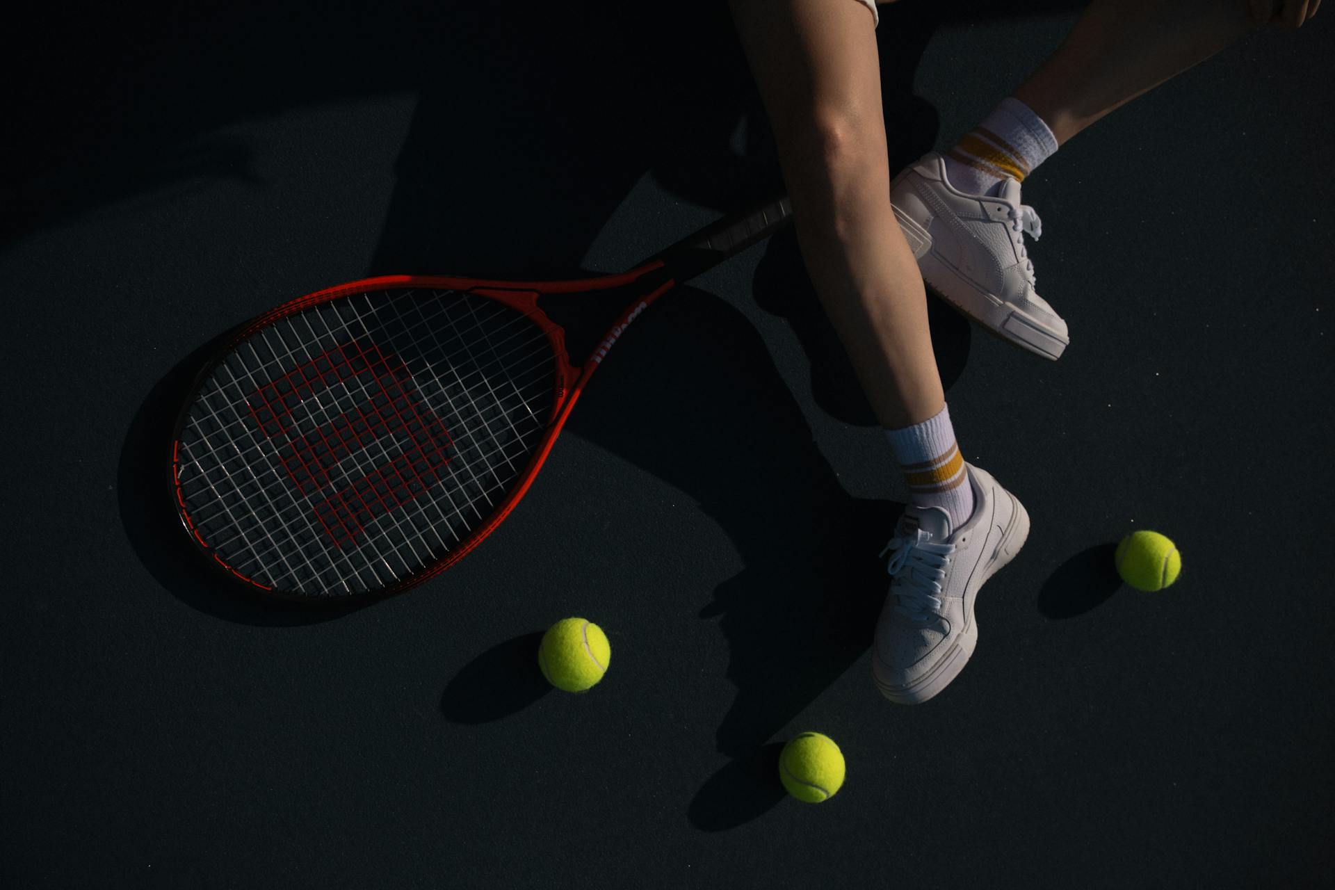 Close-up of a court with a tennis racket, three tennis balls, and a player's legs with white sneakers
