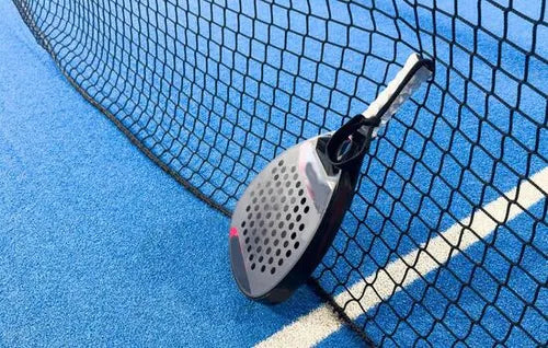 A padel racket and ball resting against the net on a blue padel court.