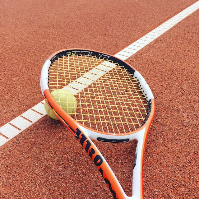 A tennis racket and tennis ball laid on a tennis court.